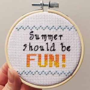 Pode incluir: Um aro de bordado branco com um padrão de ondas azul e branco na parte superior e inferior. O texto "Summer should be FUN!" está bordado em preto e laranja.