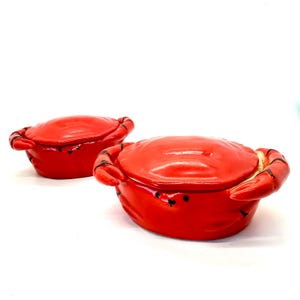 May include: Two red ceramic crab-shaped dishes with lids. The dishes have black accents on the claws and eyes. The crab dishes are on a white background.