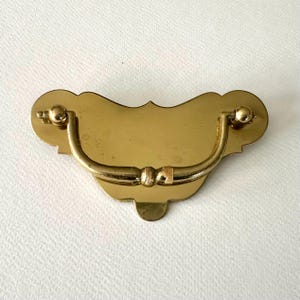 May include: A gold-toned metal drawer pull with a decorative, scalloped edge and a curved handle.