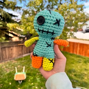 May include: A crocheted voodoo doll with a blue body, yellow and orange legs, and a black button eye. The doll has a stitched X on its chest and a needle sticking out of its head.