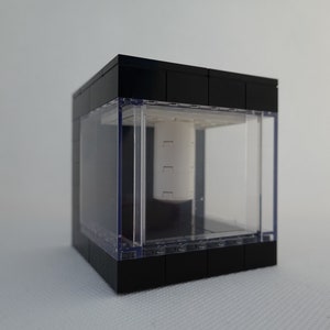 Pode incluir: Uma vitrine de exibição em forma de cubo preto com uma frente de acrílico transparente e um objeto cilíndrico branco dentro.