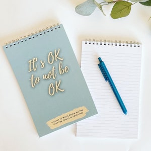 Può includere: Un quaderno a spirale blu con il testo "It's OK to not be OK" in lettere dorate. Il quaderno ha un riquadro di testo più piccolo che dice "(USE ME TO WRITE DOWN ALL THE "NOT OK THINGS IN YOUR LIFE)". Il quaderno è su una superficie bianca accanto a un quaderno a righe bianco con una penna blu.