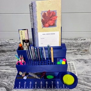 Puede incluir: Un organizador azul para suministros de ganchillo. Cuenta con un bloc de notas con un diseño de hoja roja, una variedad de ganchos de ganchillo, tijeras y una cinta métrica. El organizador tiene una regla a lo largo del borde frontal y una sección para accesorios.