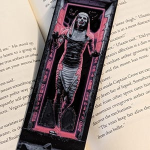 May include: A pink and black bookmark featuring a mummy-like figure with a white bandage-like outfit and a pink glow around it. The figure is standing in a doorway with hieroglyphics on the walls. The bookmark is a great way to mark your place in a book.