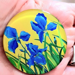 Può includere: Una spilla circolare con un vivace dipinto di tre fiori di iris blu con steli e foglie verdi su uno sfondo giallo brillante. La superficie dell'accessorio rotondo è liscia e lucida, mettendo in risalto le pennellate dettagliate dei petali.