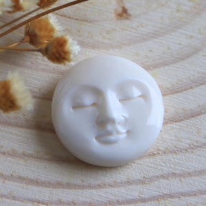 May include: A white, circular decorative piece featuring a serene face with closed eyes and a gentle smile. The piece is smooth and glossy, with subtle details. The background is a light wood grain.