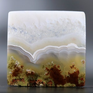 May include: Square-shaped agate stone with a landscape-like pattern. The top portion is white, transitioning to layers of beige and gray, with brown and green moss-like formations at the bottom. The stone has a polished, smooth surface.