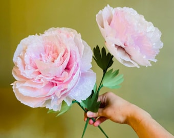 May include: Two delicate pink paper peonies with green paper leaves, held in a hand against a muted green background.