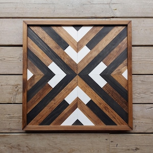 May include: Square wooden wall art with a geometric design. The artwork features a symmetrical pattern of diagonal stripes in shades of brown, black, and white, framed by a dark wood border. The piece is displayed on a wooden surface.