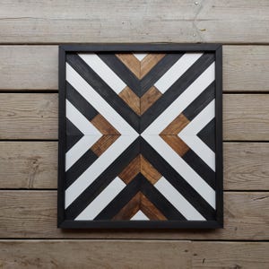 May include: A geometric wall art piece made of wood with black, white, and brown stripes. The piece is framed in black and has a modern design.