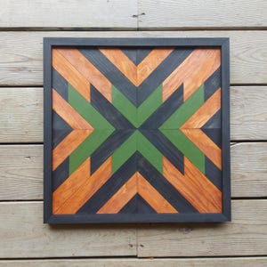 May include: Square wooden wall art in a black frame. The artwork features a geometric design with orange, green, and black chevron patterns. The wood grain is visible, adding texture to the piece. Suitable for home decor.