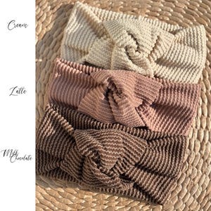 Pu&ograve; includere: Quattro fasce per capelli strutturate nei colori crema, latte e cioccolato al latte sono esposte su una superficie tessuta. Le fasce hanno un design annodato. Le parole "Cream", "Latte" e "Milk Chocolate" sono scritte in corsivo.