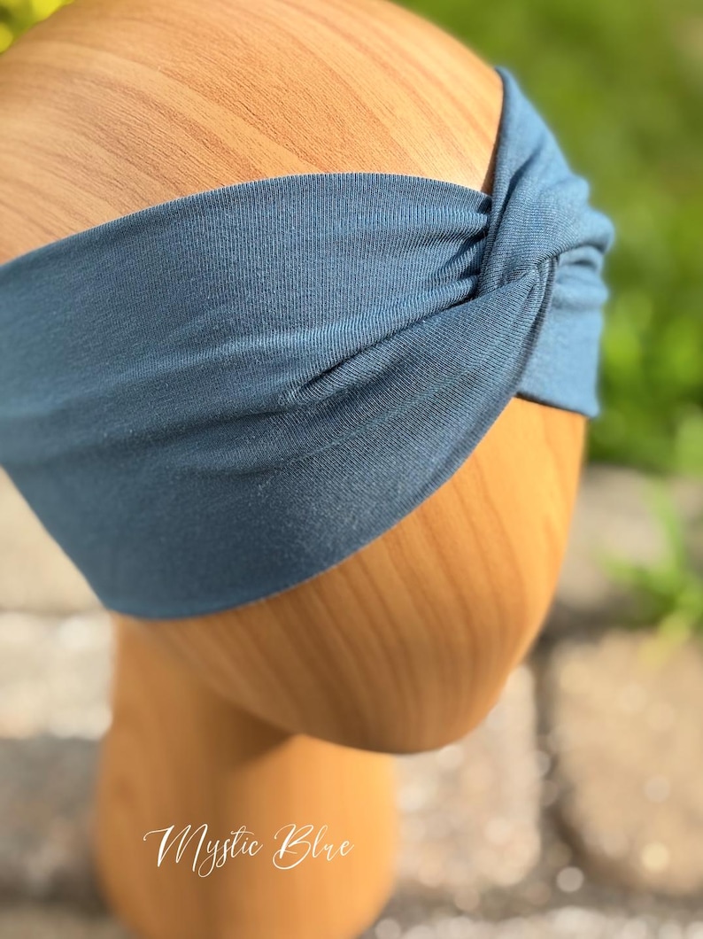 May include: A blue headband with a twisted knot design. The headband is made of a soft fabric and is displayed on a wooden head form. The text "Mystic Blue" is written in cursive at the bottom of the image.