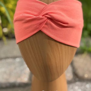 May include: Coral-colored headband with a twisted knot design. The headband is made of soft fabric and is displayed on a wooden head form. The text "Coral Pop" is written in cursive at the bottom of the image.