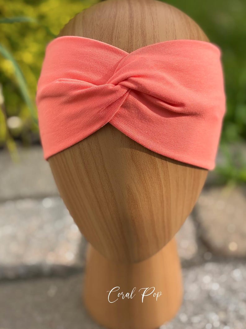 May include: Coral-colored headband with a twisted knot design. The headband is made of soft fabric and is displayed on a wooden head form. The text "Coral Pop" is written in cursive at the bottom of the image.