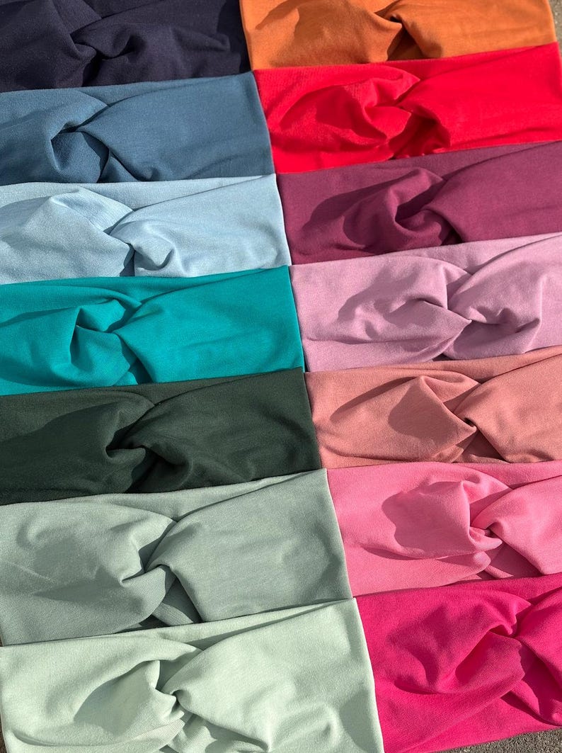 May include: A collection of colorful headbands in various shades, including navy, teal, red, and pink. Each headband features a twisted knot design, showcasing a range of solid colors. The headbands are made of soft fabric.