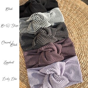Pu&ograve; includere: Una collezione di fasce per capelli annodate in vari colori, tra cui nero, a righe bianche e nere, nero antracite, melanzana e lilla. Le fasce sono realizzate in tessuto strutturato e sono disposte su una superficie intrecciata. I nomi dei colori sono scritti in corsivo.