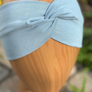 May include: A light blue headband with a twisted knot design. The headband is made of soft fabric and is displayed on a wooden head form. The text "Soft Blue" is visible at the bottom of the image.