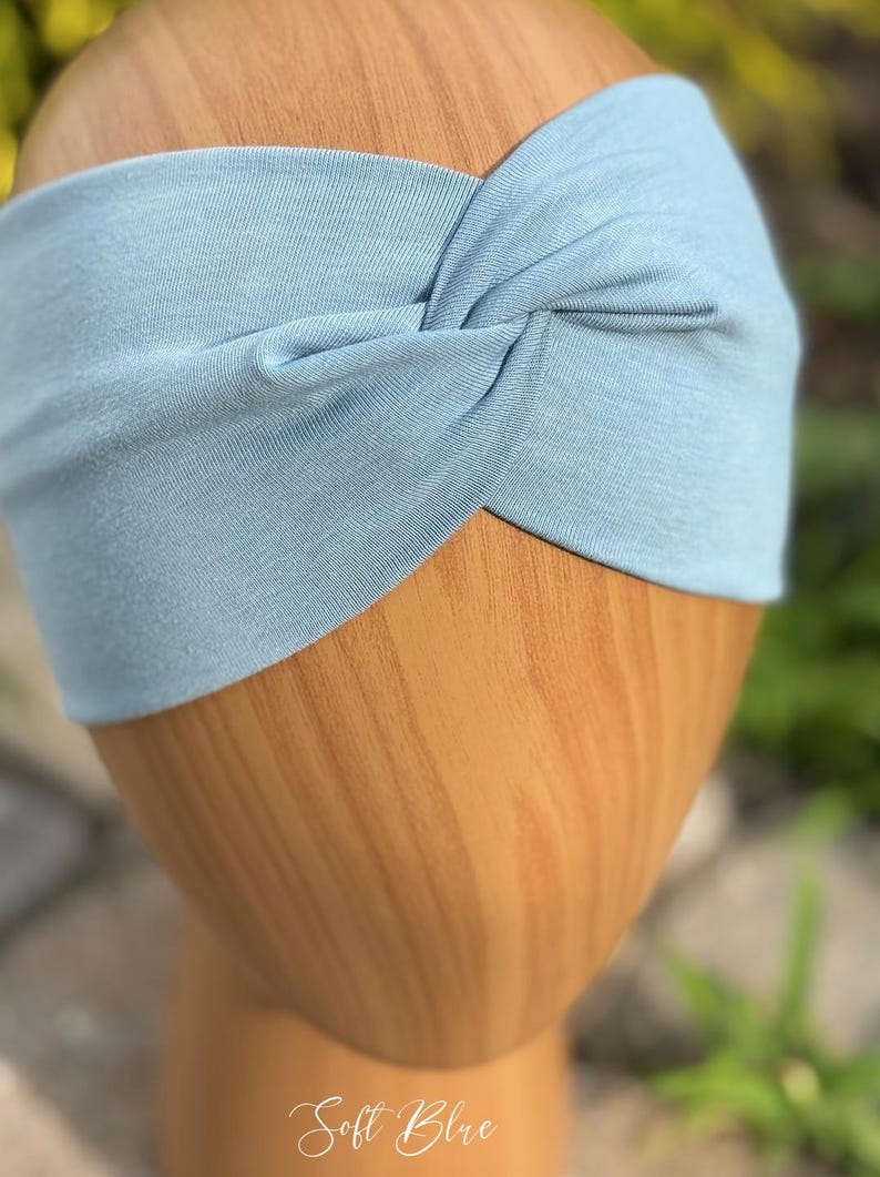 May include: A light blue headband with a twisted knot design. The headband is made of soft fabric and is displayed on a wooden head form. The text "Soft Blue" is visible at the bottom of the image.
