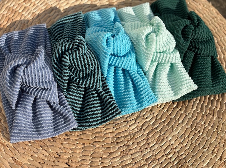 Pu&ograve; includere: Un assortimento di cinque fasce per capelli annodate in varie tonalit&agrave; di blu e verde. Le fasce hanno un motivo strutturato a forma di waffle e sono disposte su una superficie intrecciata marrone chiaro. I colori includono azzurro, turchese e verde scuro.