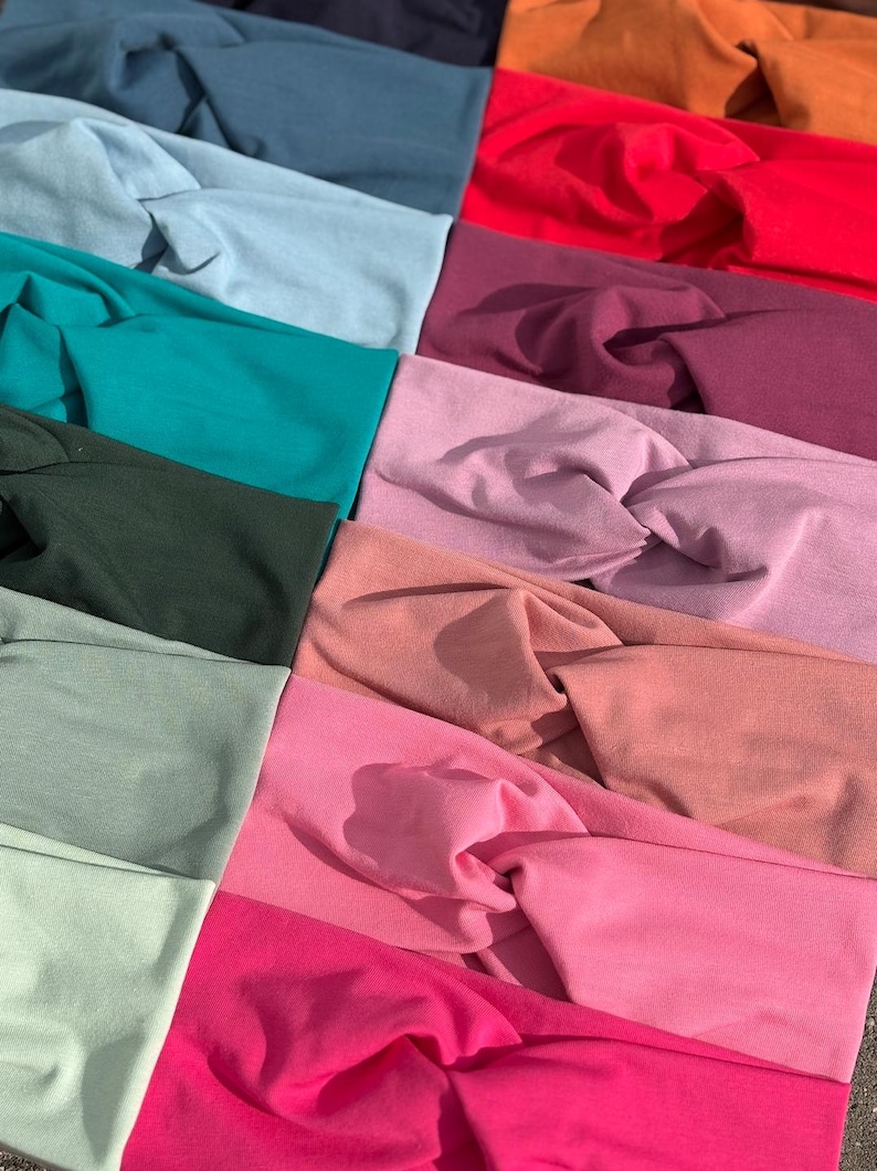 May include: A collection of colorful headbands in various shades, including teal, pink, red, and green. The headbands are made of soft fabric and feature a twisted knot design. The assortment showcases a range of colors, from light pastels to vibrant hues.