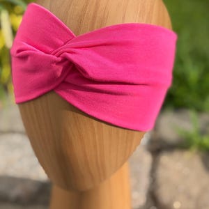 May include: A bright pink headband with a twisted knot design. The headband is displayed on a wooden head form. The text "Party Pink" is written in cursive at the bottom of the image. This accessory is suitable for various hairstyles.