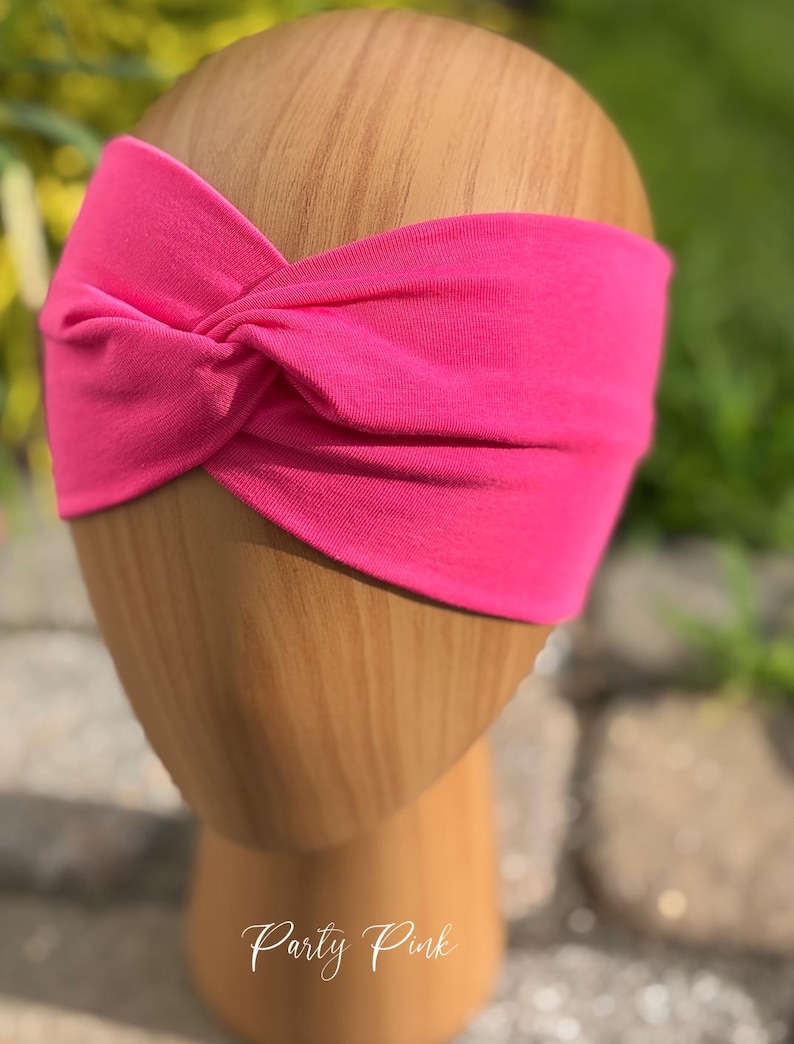 May include: A bright pink headband with a twisted knot design. The headband is displayed on a wooden head form. The text "Party Pink" is written in cursive at the bottom of the image. This accessory is suitable for various hairstyles.