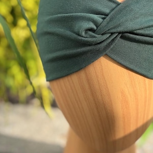 May include: A dark hunter green headband with a twisted knot design. The headband is made of a soft fabric and is displayed on a wooden head form. The text "Hunter Green" is written in cursive at the bottom of the image.