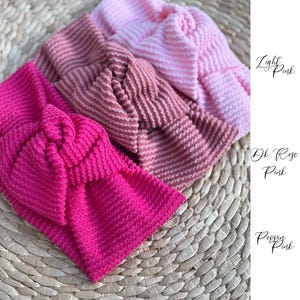 Pu&ograve; includere: Tre fasce per capelli rosa in diverse tonalit&agrave;: rosa chiaro, rosa rosa scuro e rosa papavero. Ogni fascia presenta un tessuto strutturato e a coste e un design annodato. Le fasce sono esposte su una superficie intrecciata marrone chiaro.