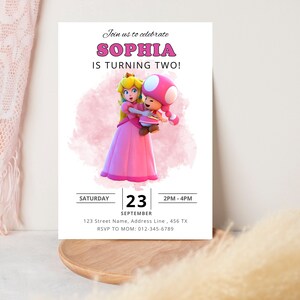 Puede incluir: Una invitaci&oacute;n de cumplea&ntilde;os rosa y blanca con una imagen de dibujos animados de la princesa Peach y Toad de la serie de videojuegos Super Mario Bros. La invitaci&oacute;n dice "&iexcl;&Uacute;nete a nosotros para celebrar que Sophia cumple dos a&ntilde;os!" e incluye la fecha, la hora y la informaci&oacute;n de RSVP.