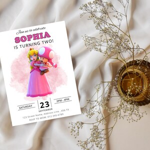 Puede incluir: Una invitaci&oacute;n de cumplea&ntilde;os rosa y blanca con una imagen de dibujos animados de la princesa Peach y un personaje de Toad. La invitaci&oacute;n dice "&iexcl;&Uacute;nete a nosotros para celebrar que Sophia cumple dos a&ntilde;os!" e incluye la fecha, la hora y la informaci&oacute;n de RSVP.