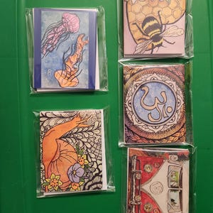 May include: A collection of five small, colorful art prints in clear plastic packaging. The prints feature various illustrations, including jellyfish, a bee on a honeycomb, a hand with flowers, an abstract design, and a vintage vehicle. The prints are displayed on a green surface.