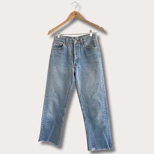 May include: Light blue denim jeans featuring a high-rise waist, a button-fly closure, and straight legs with raw, frayed hems. The jeans have a classic five-pocket design with copper-toned rivets and are displayed on a wooden hanger.