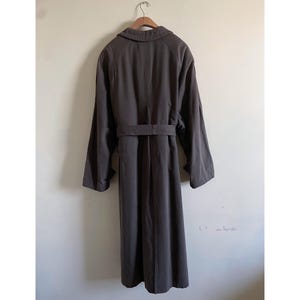 Puede incluir: Una gabardina larga de color marr&oacute;n oscuro con cintur&oacute;n y cuello. La gabardina est&aacute; colgada de una percha de madera.