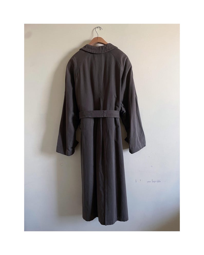 Puede incluir: Una gabardina larga de color marr&oacute;n oscuro con cintur&oacute;n y cuello. La gabardina est&aacute; colgada de una percha de madera.
