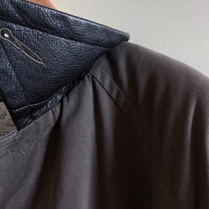 Puede incluir: Una chaqueta gris oscuro con cuello de cuero negro. El cuello tiene un detalle cosido.