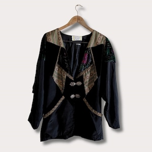 May include: Black vintage jacket with decorative trim and ornate buttons. The jacket features a V-neck design with patterned fabric accents in shades of gold, green, and pink. The long-sleeved jacket is hanging on a wooden hanger.