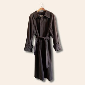 Puede incluir: Una gabardina larga de color marr&oacute;n oscuro con cintur&oacute;n y cierre de botones. La gabardina tiene un dise&ntilde;o cl&aacute;sico y est&aacute; hecha de un tejido duradero.