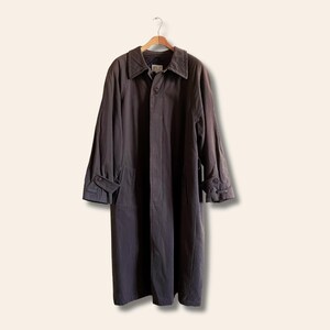 Puede incluir: Una gabardina larga de color gris oscuro con cierre de botones y cuello. La gabardina tiene un bolsillo en el pecho y dos bolsillos con solapa en la parte delantera inferior.