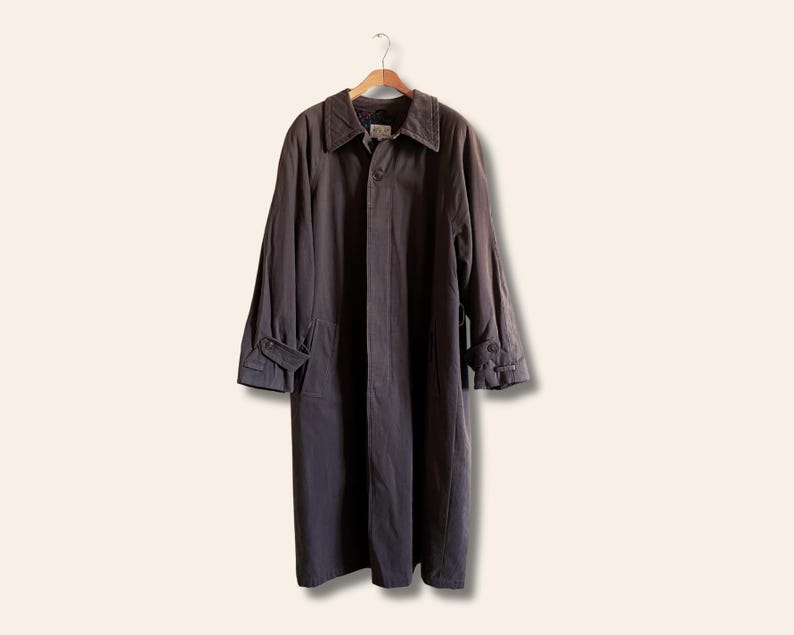 Puede incluir: Una gabardina larga de color gris oscuro con cierre de botones y cuello. La gabardina tiene un bolsillo en el pecho y dos bolsillos con solapa en la parte delantera inferior.