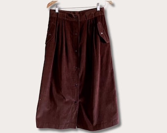 May include: A long, button-down skirt in a deep burgundy color. The skirt features pleats, two front pockets, and is made of a soft, corduroy-like material. It hangs on a wooden hanger against a neutral background.
