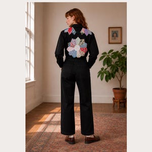 May include: A black long-sleeved jumpsuit featuring a large, multi-colored floral quilt-style patch on the back and smaller matching patches on the shoulders. The garment has a collared neckline, long sleeves, and straight-leg trousers.