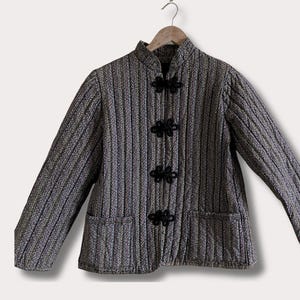 May include: A quilted jacket with a mandarin collar and long sleeves. The jacket features a vertical striped pattern in shades of gray and black, with three black frog closures down the front. Two pockets are visible.