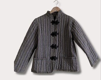 May include: A quilted jacket with a mandarin collar and long sleeves. The jacket features a vertical striped pattern in shades of gray and black, with three black frog closures down the front. Two pockets are visible.