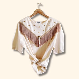 May include: A white cotton shirt with a brown fringe trim and gold accents. The shirt has a knotted front and a relaxed fit.