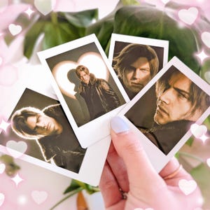 May include: Four instant photos featuring a man in a black leather jacket. One photo has a heart-shaped light behind the man. The photos are held in a hand with light blue nail polish, with a background of green leaves and pink heart and star decorations.