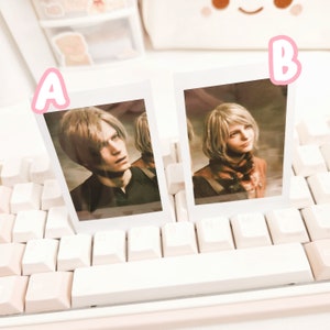 May include: Two Polaroid-style photo frames with white borders, each featuring a portrait of a person. The frame on the left shows a person with short, light brown hair, wearing a black shirt. The frame on the right shows a person with long, light brown hair, wearing a brown shirt. Both frames are placed on a white keyboard.