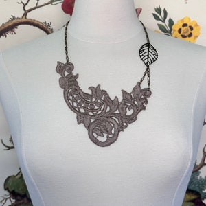 May include: A gray lace necklace with an ornate floral design. The necklace has a delicate, openwork pattern and a leaf-shaped pendant on a chain. The necklace is displayed on a white mannequin.