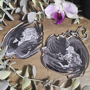 Può includere: Due portachiavi in acrilico con chiusura a forma di cuore. Ogni portachiavi presenta un'illustrazione in bianco e nero di una figura alata che ne abbraccia un'altra. I portachiavi sono appoggiati su una superficie di legno con verde e un'orchidea bianca e viola.