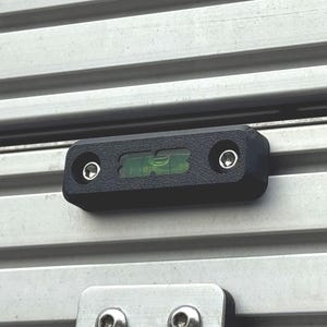 May include: A black rectangular spirit level with a green liquid vial and two silver screws, mounted horizontally onto a silver aluminum extrusion profile. Below the level is a silver metal hinge secured with two additional silver screws.